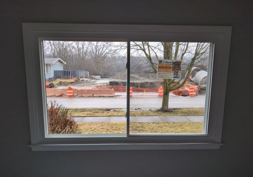 View through a window showing a street with orange construction barriers, a tree, a sidewalk, and construction equipment in the background.