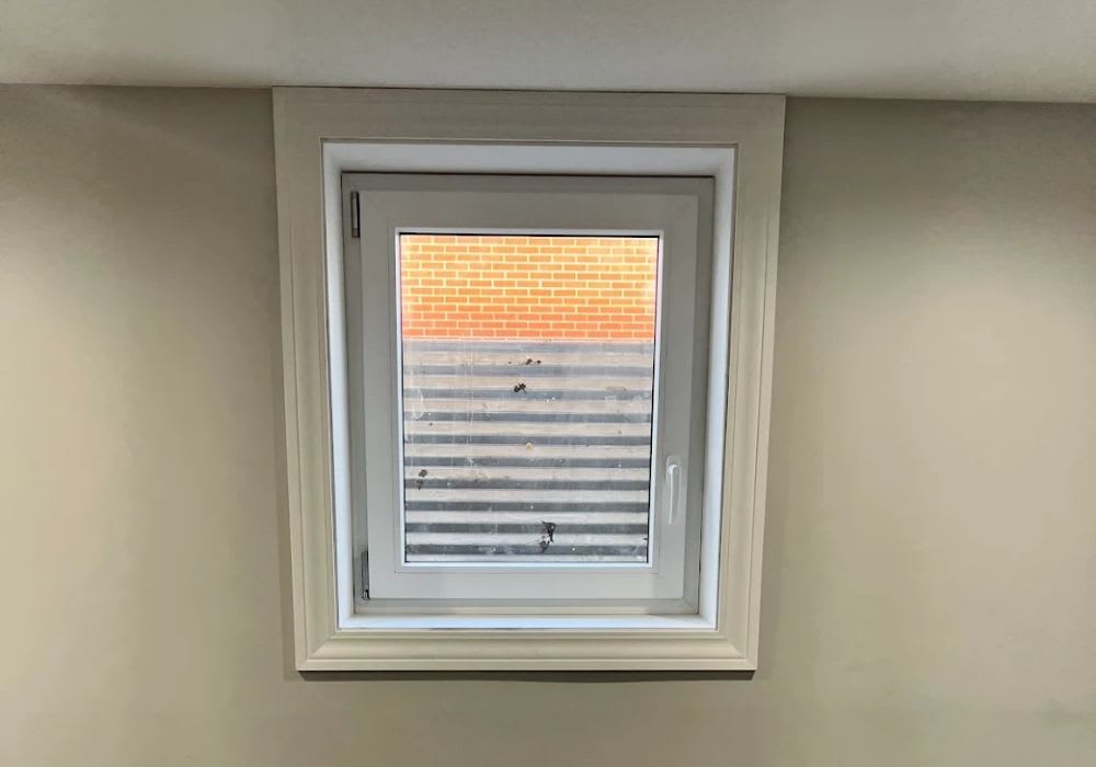 A small white-framed window set in a beige wall, looking out onto a corrugated metal window well and a brick wall.