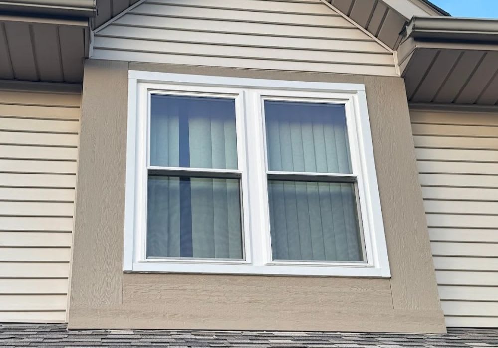 A double-hung window with closed blinds is set in a beige exterior wall beneath the roofline of a house with horizontal siding.
