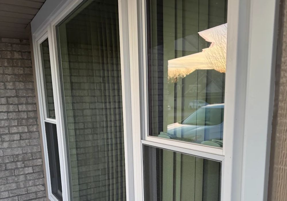A row of house windows, one with a large crack in the upper pane, reflecting a car and sunset outside.