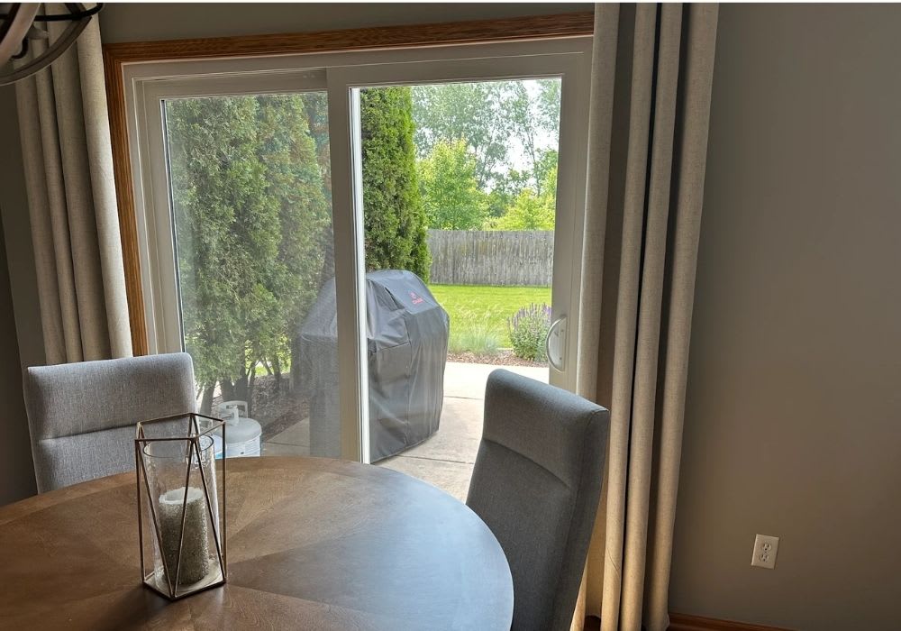 A dining area with a round wooden table and two gray chairs is next to a sliding glass door that opens to a patio with a covered grill and green backyard.