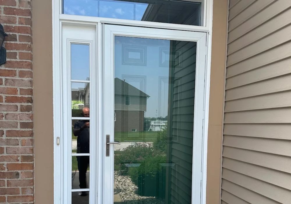 A white front door with a glass storm door, surrounded by tan siding and red brick, with a reflection of a person taking the photo visible in the glass.