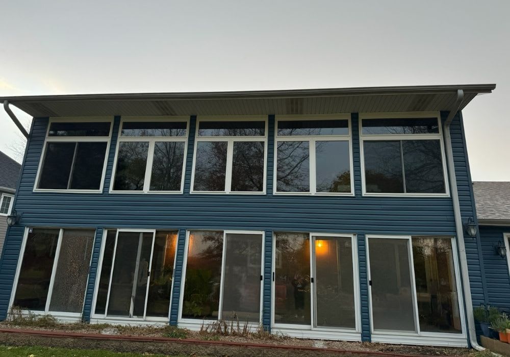 Two-story house with blue siding and large windows; upper floor has multiple rectangular windows, lower floor has several sliding glass doors.