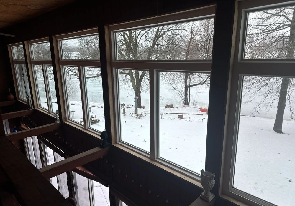 Large windows overlook a snowy landscape with bare trees, a frozen body of water, and scattered outdoor objects visible outside.