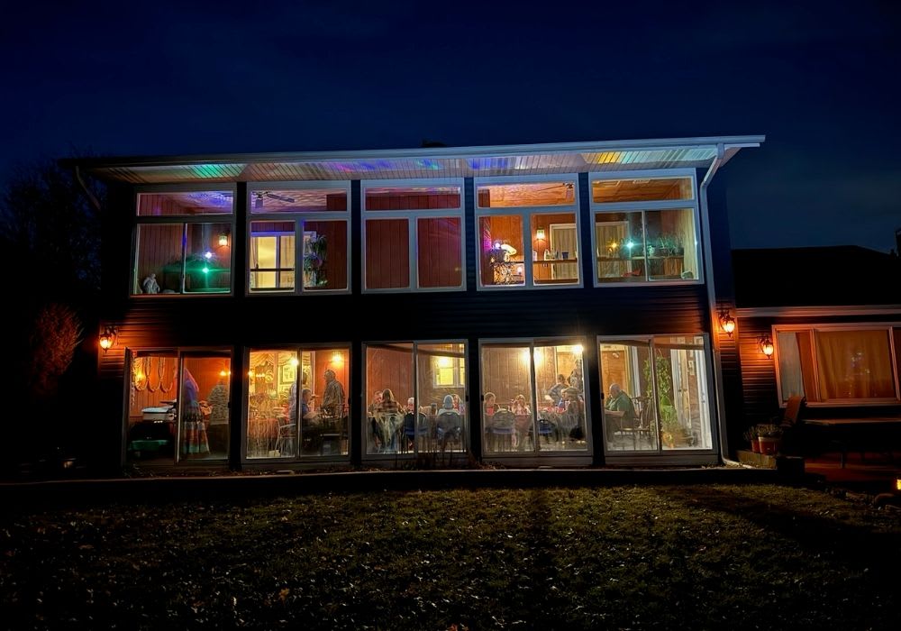 A two-story house at night with people sitting and dining inside a brightly lit room; colorful lights are visible on the upper floor.