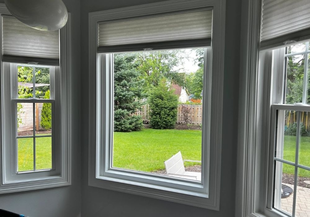 Large windows overlook a green backyard with grass, trees, shrubs, a wooden fence, and a white chair visible outside.