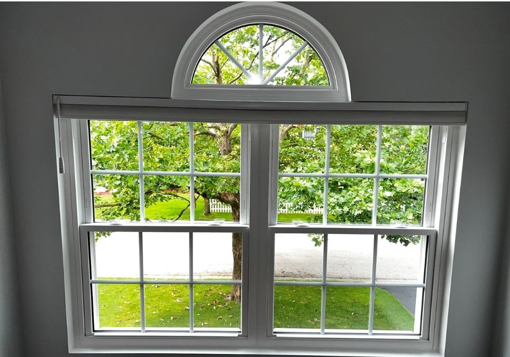A large window with multiple panes and an arched top shows a view of a leafy tree, a street, and a grass lawn outside.