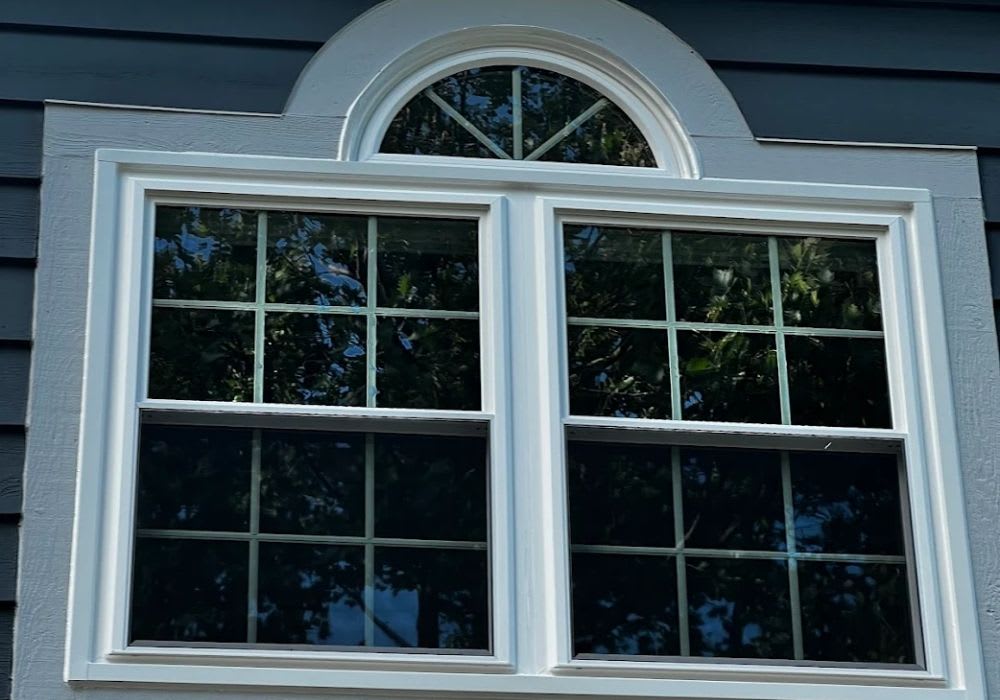 A house window with white trim and divided panes, topped by a half-round window, reflects trees and sky. The exterior wall is painted dark gray with horizontal siding.
