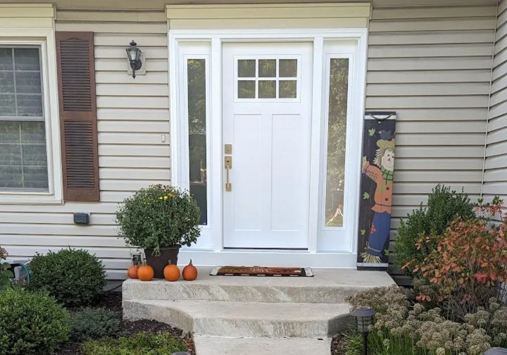 A white front door with side windows, three small pumpkins, a potted plant, and a scarecrow banner decorate the entrance of a beige house with shrubs and flowers.