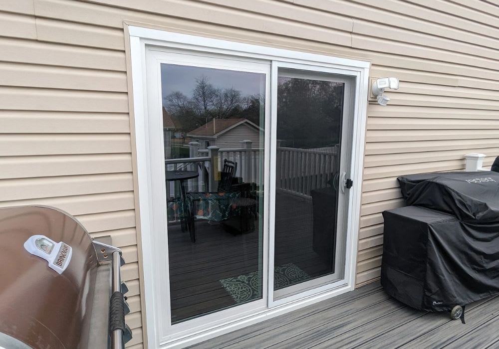 A closed sliding glass door on a house with tan siding, leading to a deck with a grill, covered grill, and outdoor furniture visible through the glass.