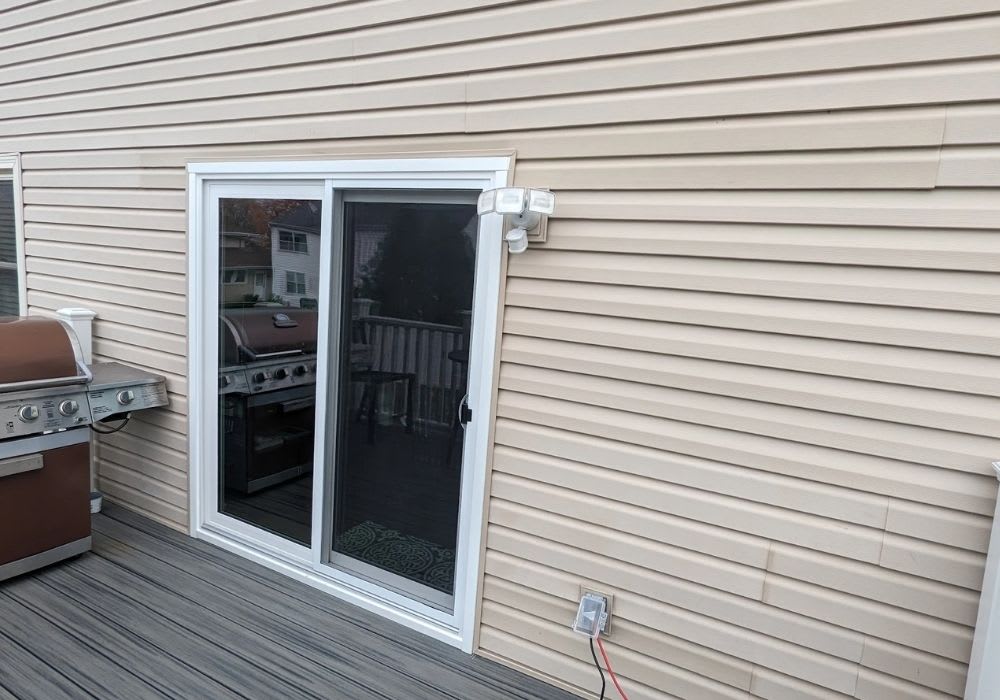 A sliding glass door is installed on a beige vinyl-sided wall, with a grill on the left and an outdoor security light and electrical outlet nearby.