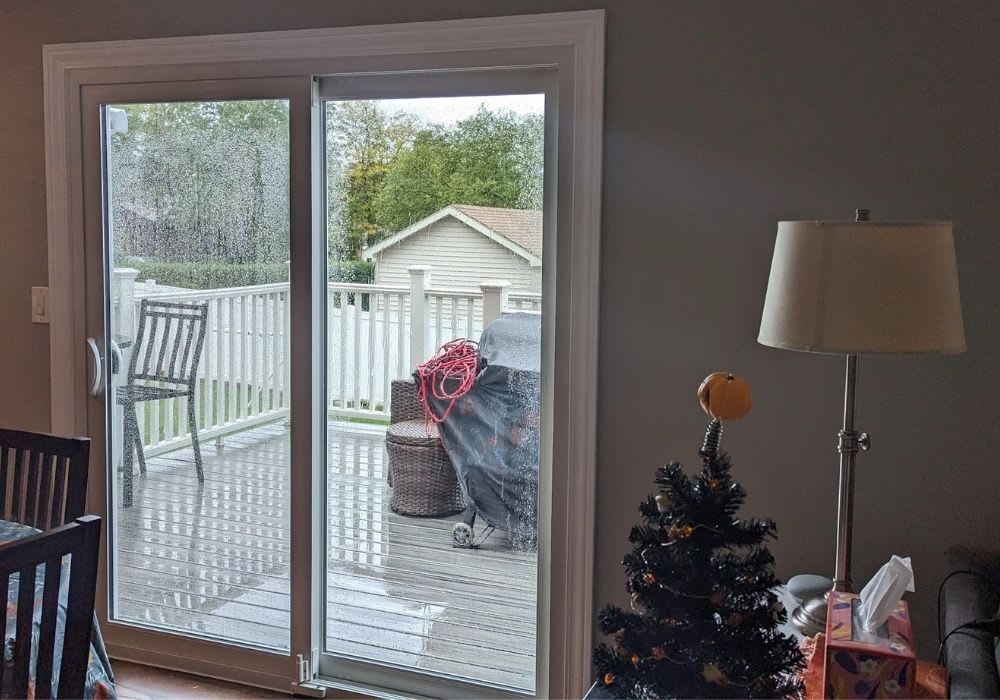 Sliding glass door with rain droplets, leading to a deck with chairs, a grill, and storage bins; inside, a lamp and a small decorated Christmas tree are visible.