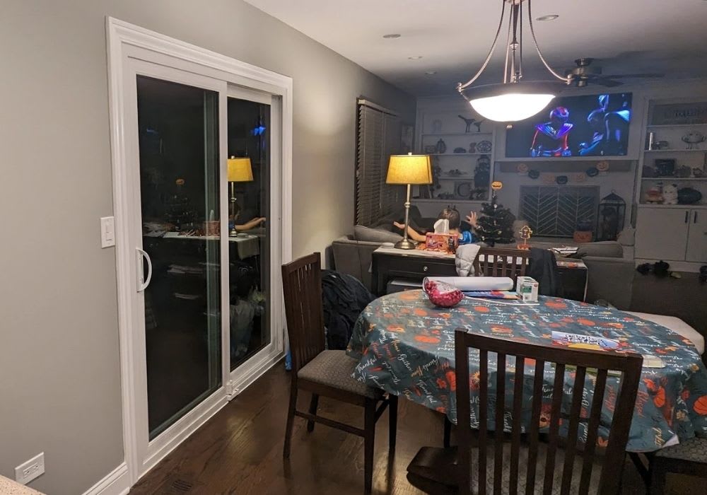 A dining area with a table and chairs is in the foreground; in the background, a person sits on a couch watching TV in a living room.