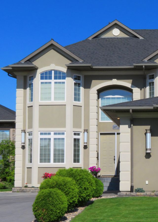 Two-story beige house with white trim, large front windows, black roof, double front door, and manicured bushes in front yard under clear blue sky.