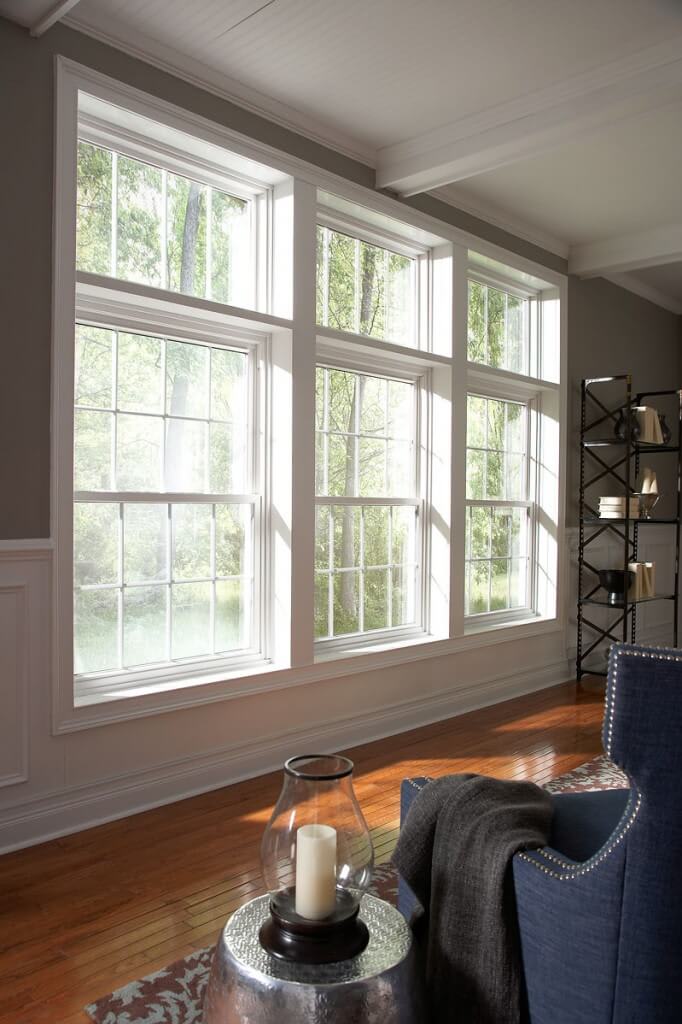 Large grid-style windows let sunlight into a living room with hardwood floors, a blue armchair, a blanket, and a small round table with a candle.