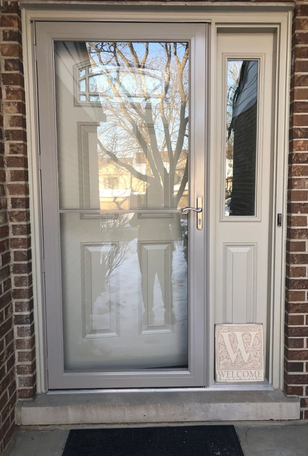 A glass storm door, installed by expert door contractors, shows a reflection of a person taking the photo, framed by brick walls, with a "Welcome" sign and black doormat at the entrance. Door installation in Chicago