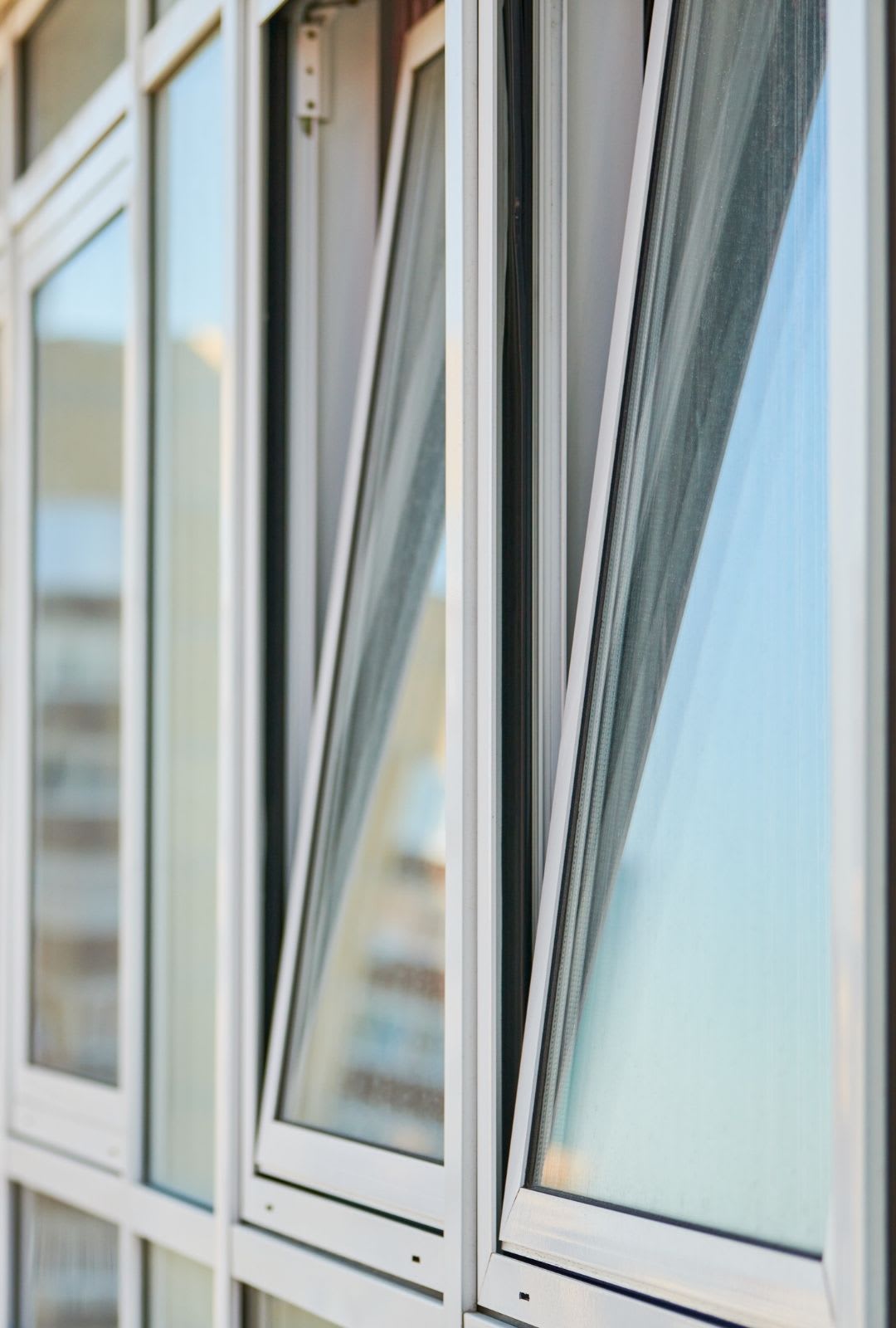 Several modern glass windows on a building facade, some of which are partially open as awning windows, reflecting the sky and nearby structures—showcasing quality window installation.