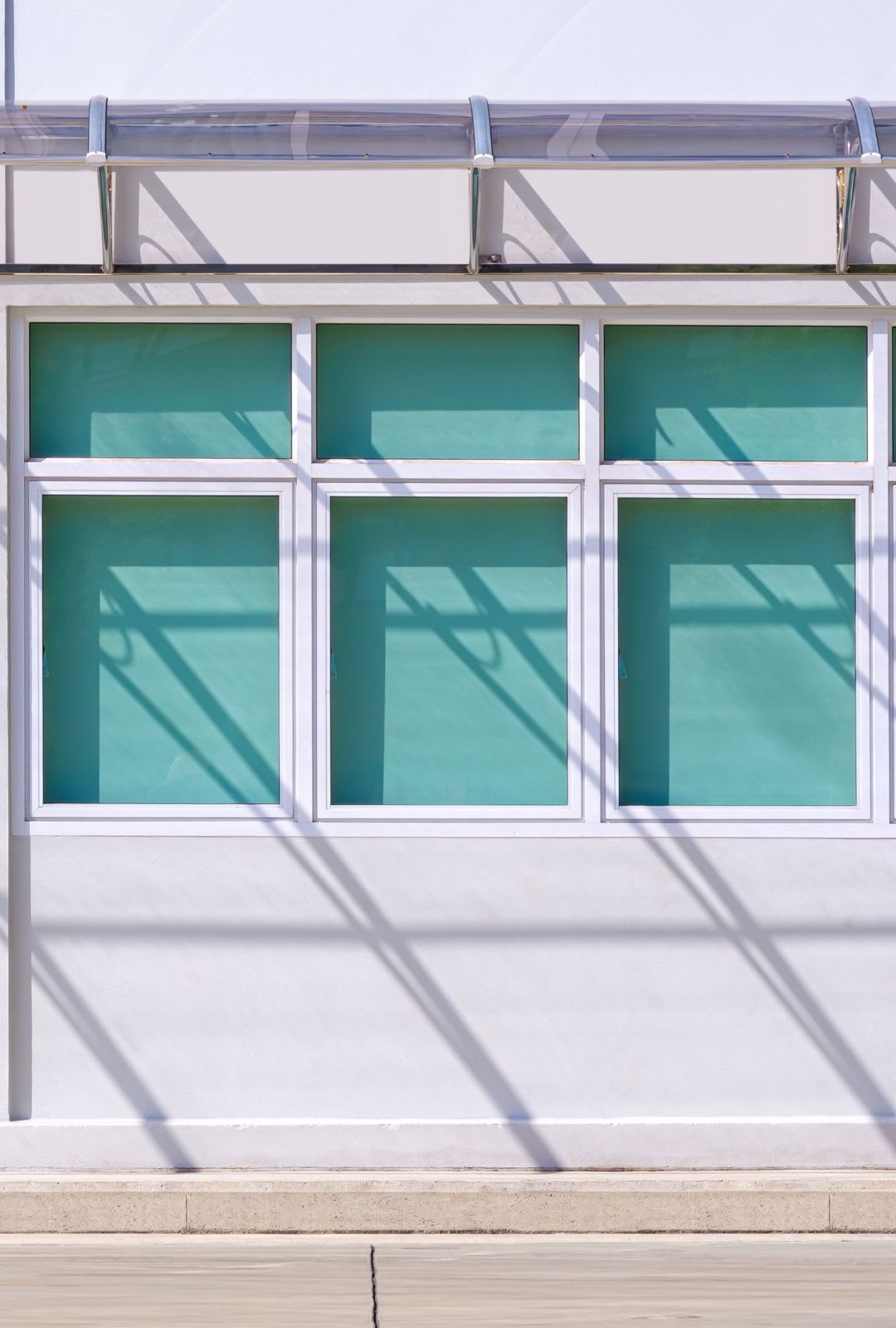 A white building with five rectangular turquoise-tinted windows and a clear awning casting shadows on the wall, showcasing sleek replacement awning windows for a modern touch.