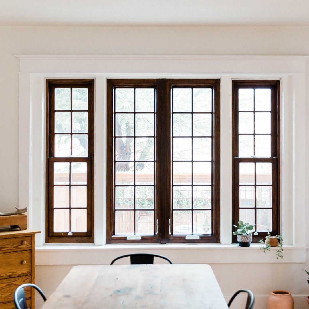 A wooden dining table and chairs sit in front of large, grid-paned windows with dark frames; custom window installation by Chicago Window Guys enhances the space. Potted plants and a chest of drawers are nearby.