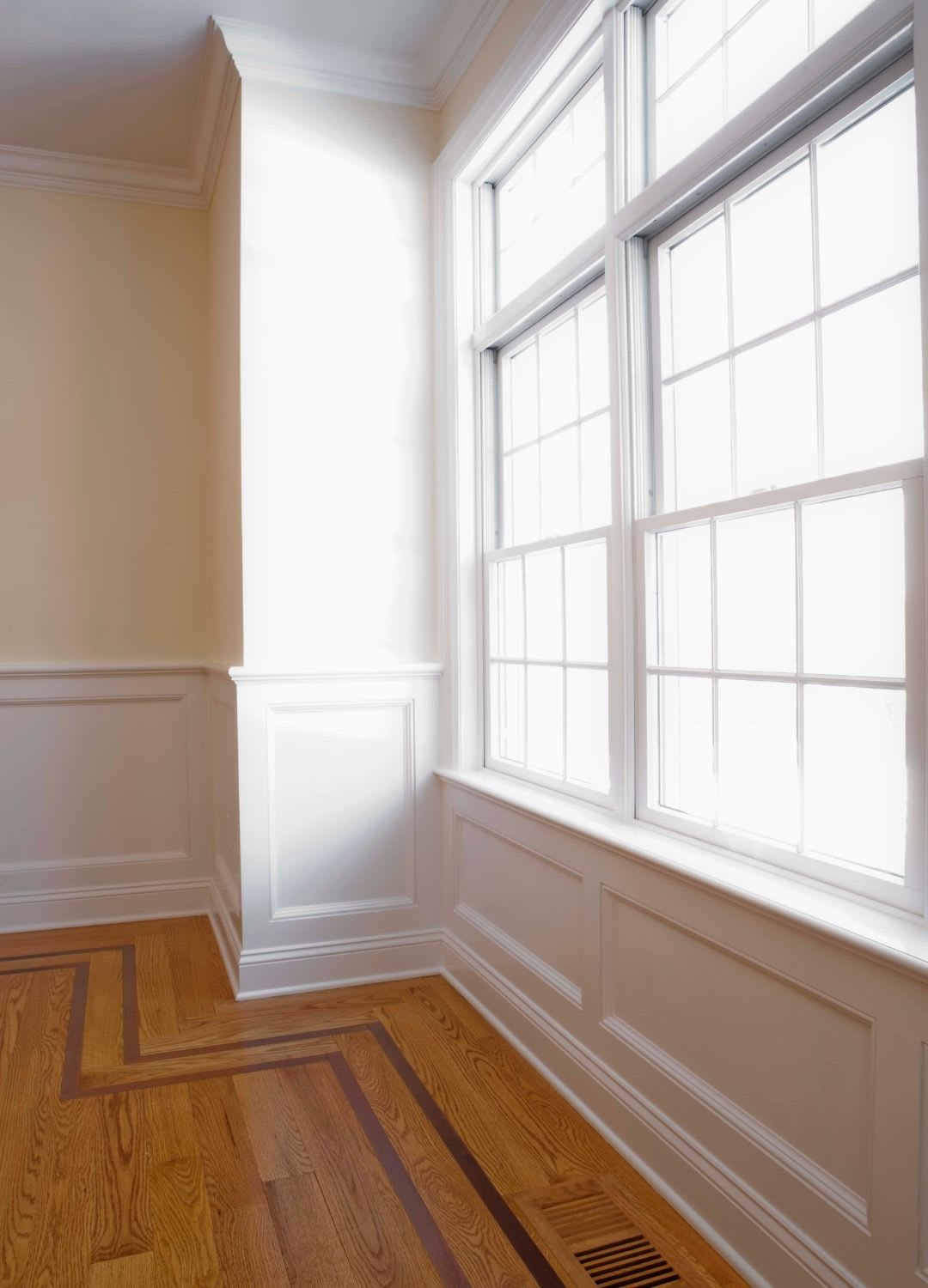 Bright sunlight streams through large, white-trimmed windows—thanks to expert custom window installation by Chicago Window Guys—onto wood flooring with decorative inlay in an empty room with cream-colored walls and wainscoting.