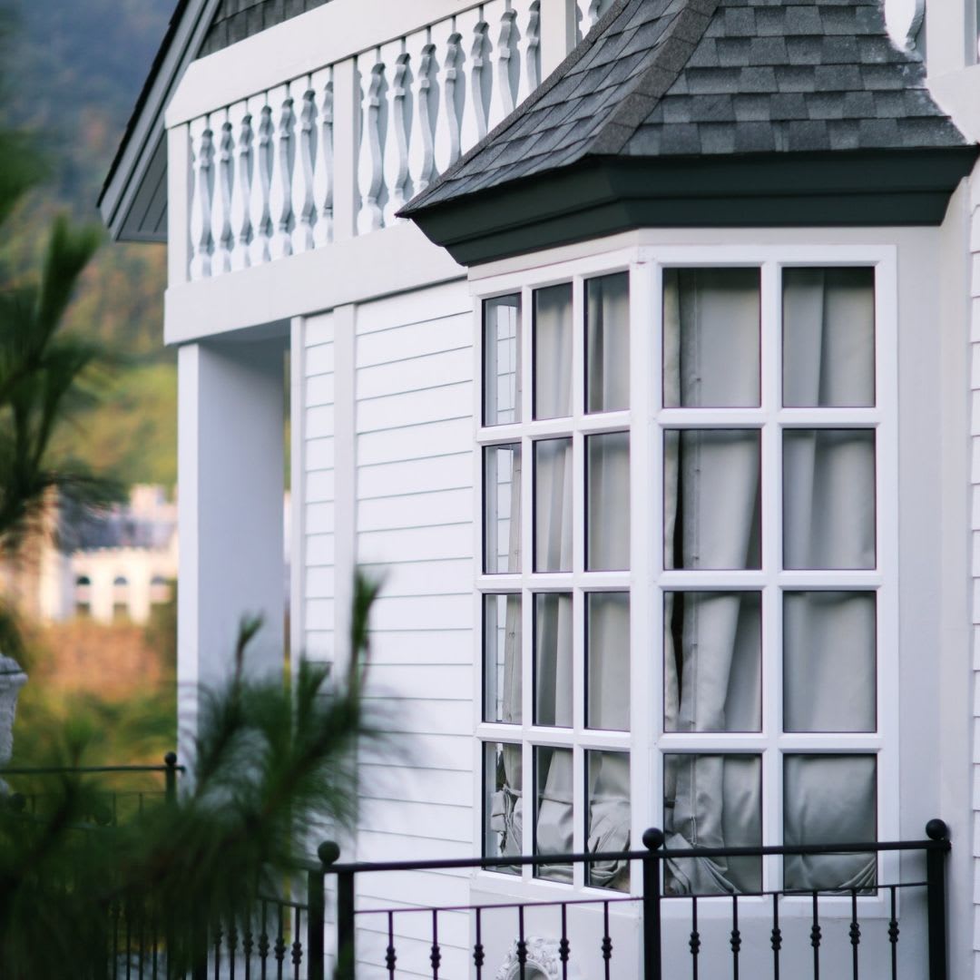 A close-up view of a white house with large bay windows—perfect for showcasing expert bay window installation by Chicago Window Guys. White curtains and a decorative balcony railing complement the lush greenery in the foreground.