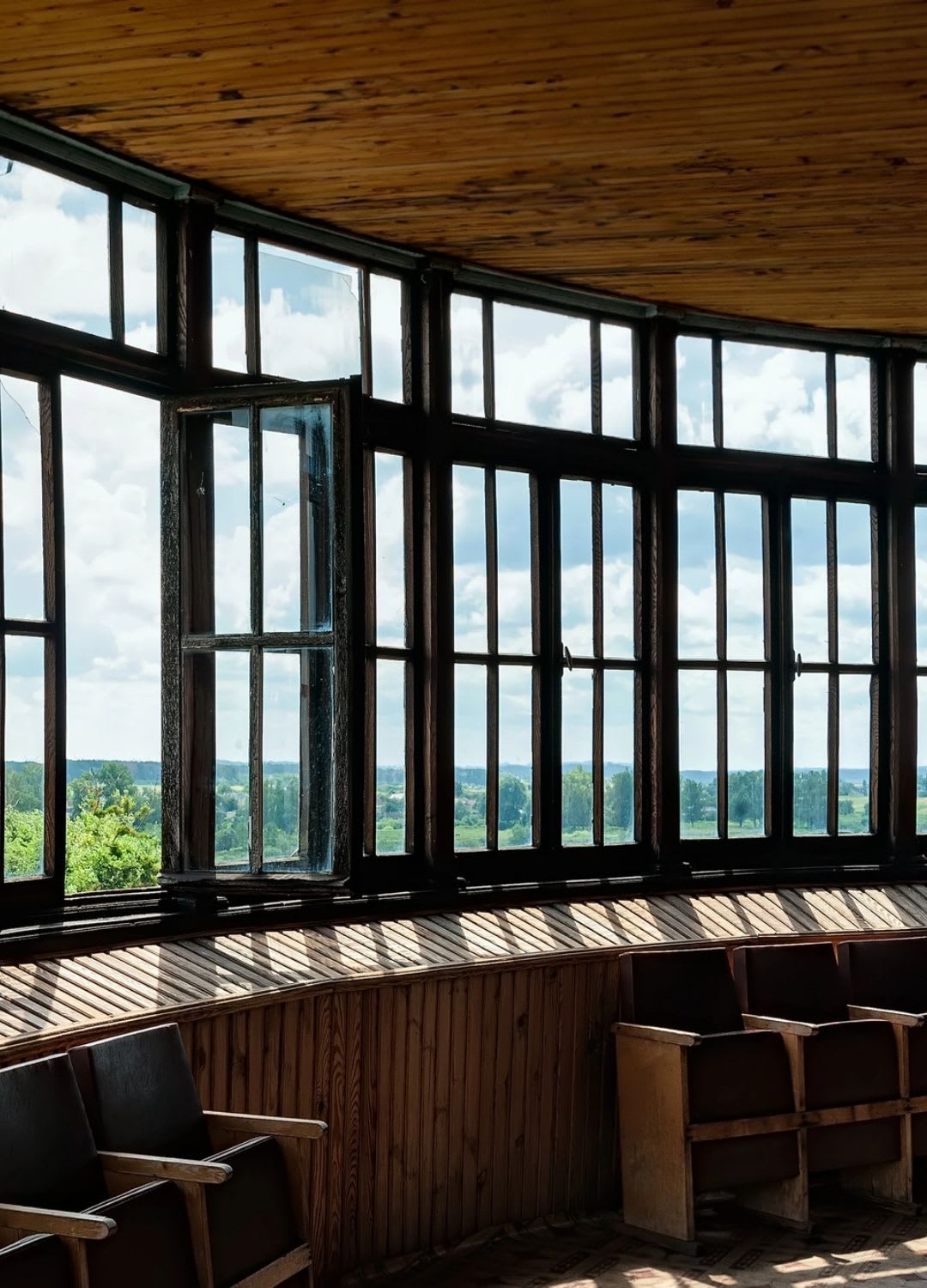 Rows of wooden chairs face large, curved replacement bow windows, one open to let in light and views of a green landscape under a partly cloudy sky.