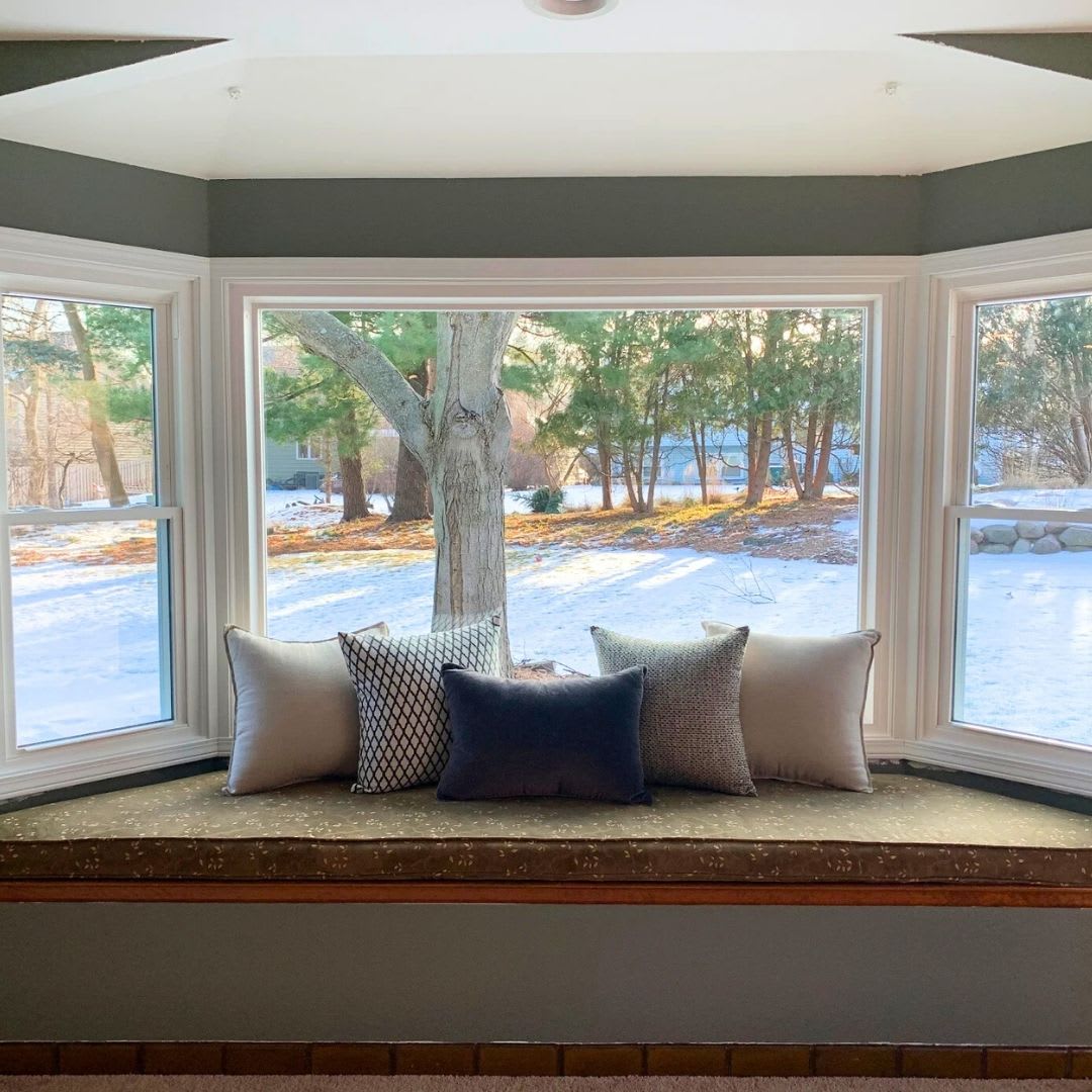 A window seat with five cushions overlooks a snow-covered yard and trees through a large bay window—perfect inspiration if you're looking to replace your bay window. Trust Chicago Window Guys for expert window replacement.