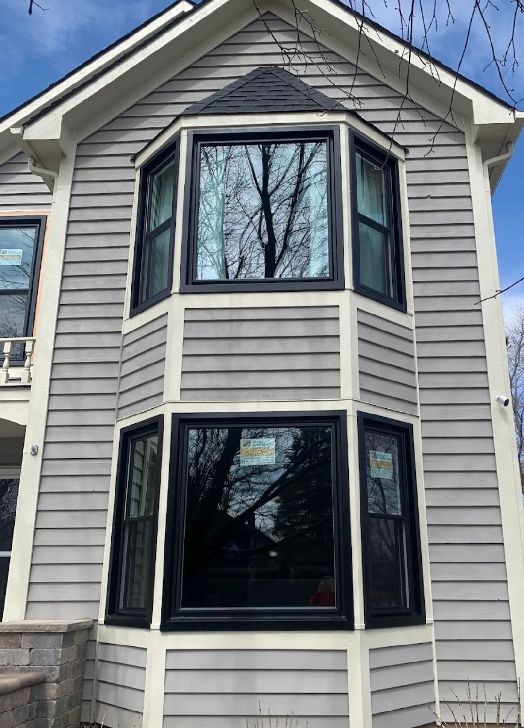 A two-story house features a light gray exterior with black-trimmed bay windows on both floors, showcasing window replacement by Chicago Window Guys and reflecting nearby trees in the glass.