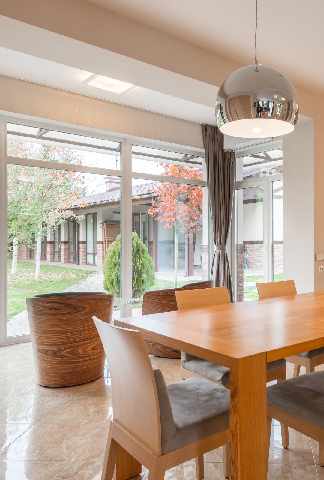 Modern dining area with a wooden table, light-colored chairs, round wooden armchairs, and a metallic pendant lamp, featuring replacement picture windows that overlook a garden filled with autumn trees.