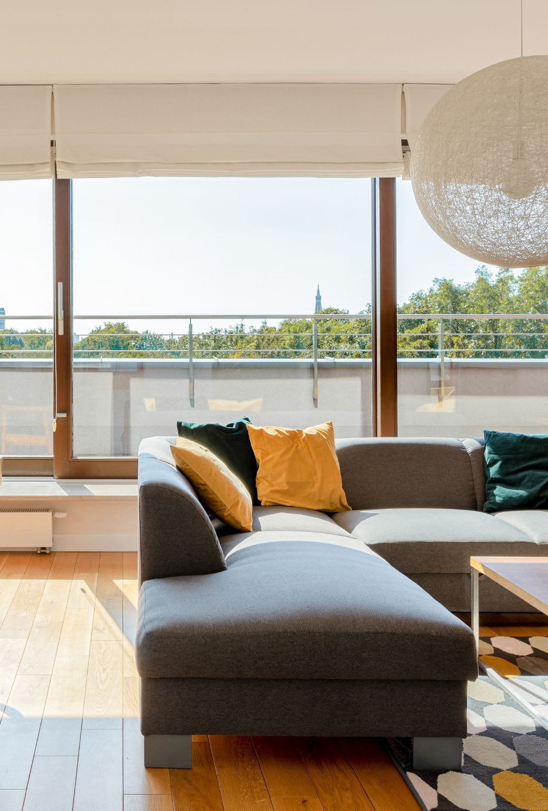 A modern living room with a gray sectional sofa, colorful pillows, wood flooring, and large custom picture window offering a balcony view of trees and a distant clock tower.