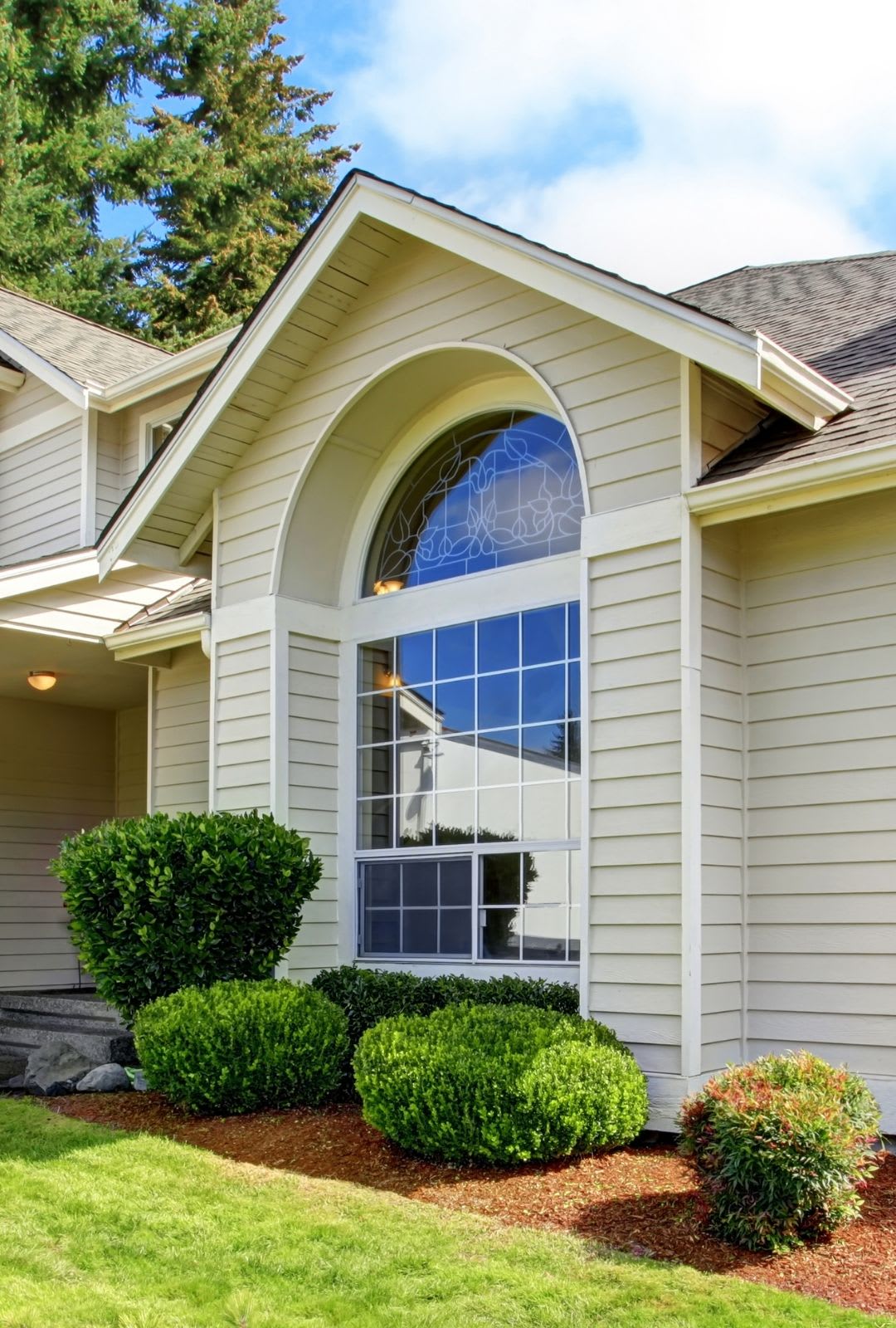 Large arched custom architectural windows with decorative glass grace the beige exterior of this house, surrounded by neatly trimmed bushes and a green lawn.