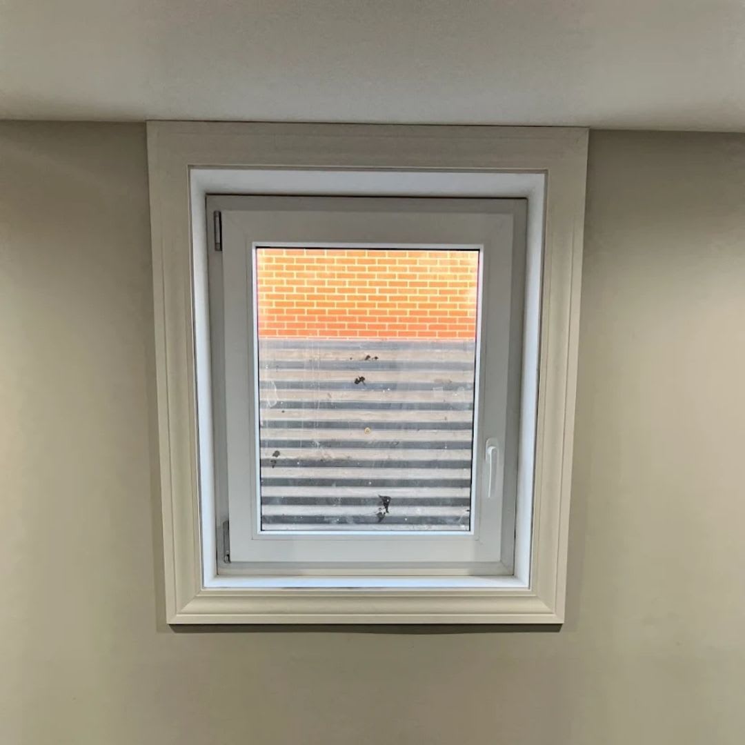 A small square basement egress window set in a beige wall, showing a view of a metal window well and a brick wall outside—perfect for those seeking basement egress window installation or replacement.