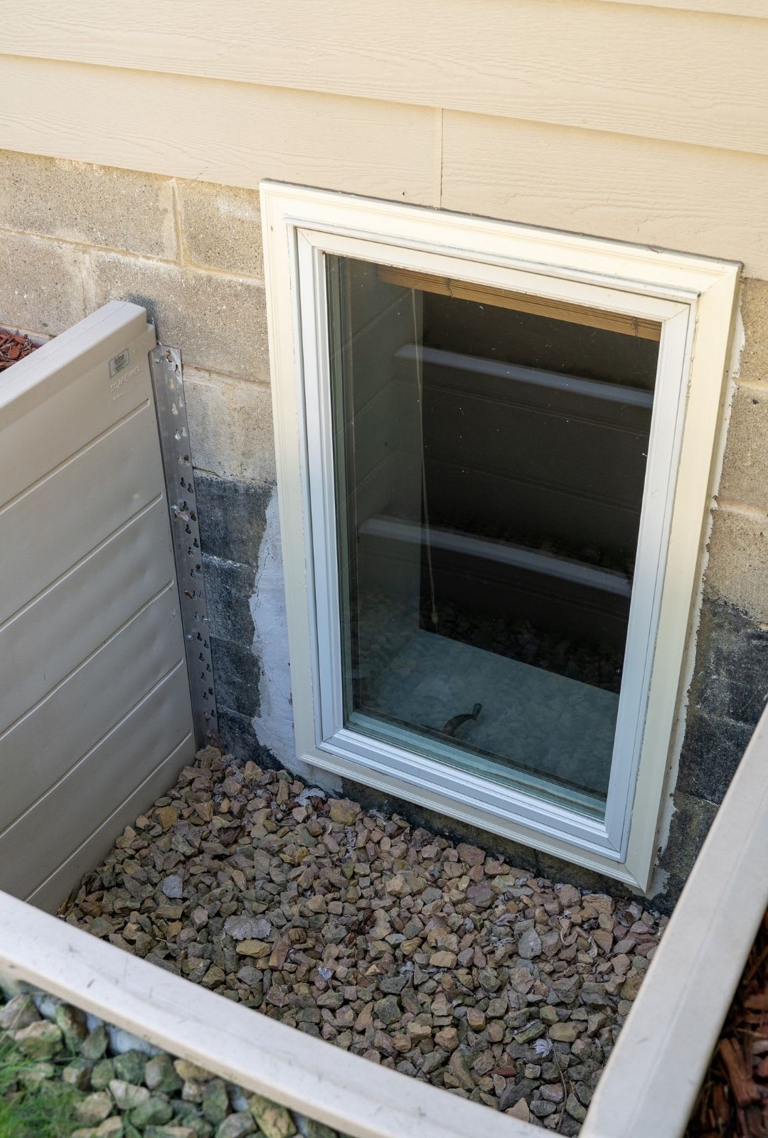 A basement window with a white frame is set into a concrete foundation wall, surrounded by a rock-filled window well—an example of professional egress window installation for safety and added natural light.