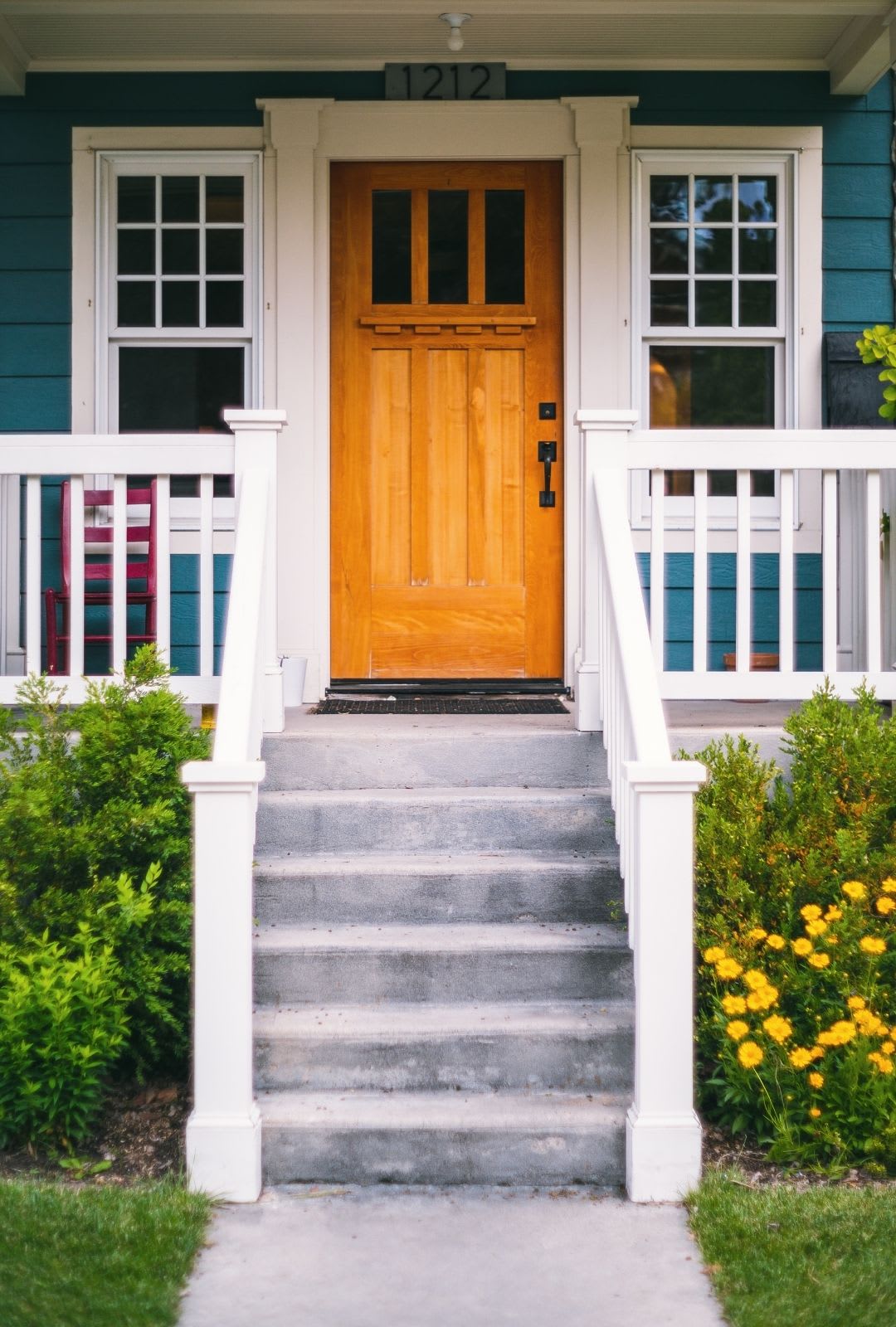 A front view of a house with blue siding, a wooden entry door replacement, white trim and railings, concrete steps, and yellow flowers along the walkway.