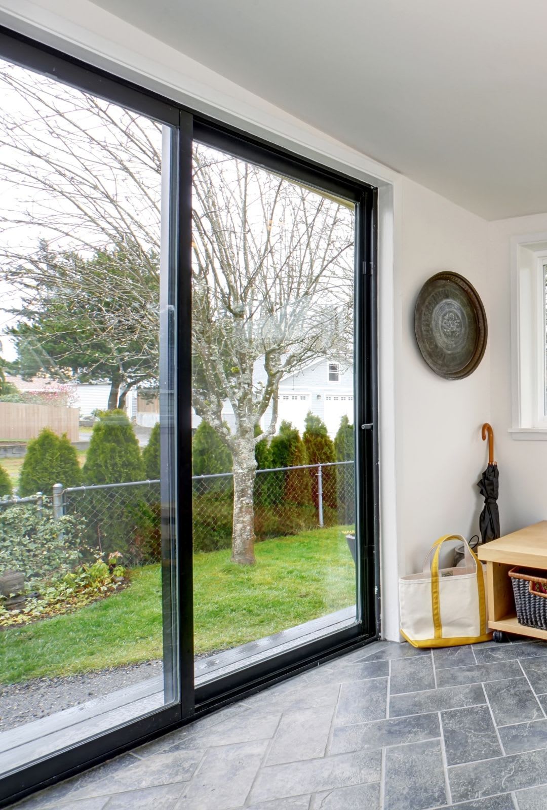 A sliding glass door by Sliding Doors Chicago opens to a backyard with a tree, green lawn, and fence; inside, tiled floor, bench, bag, umbrella, and wall decor are visible.