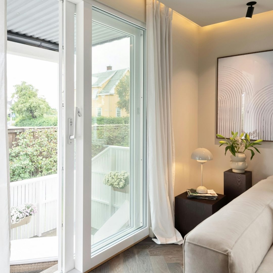 A modern living room features a sliding glass door open to a patio, white curtains, beige sofa, side table with lamp, plant, and framed artwork—perfect inspiration for those seeking sliding door replacement or ideas in Chicago.