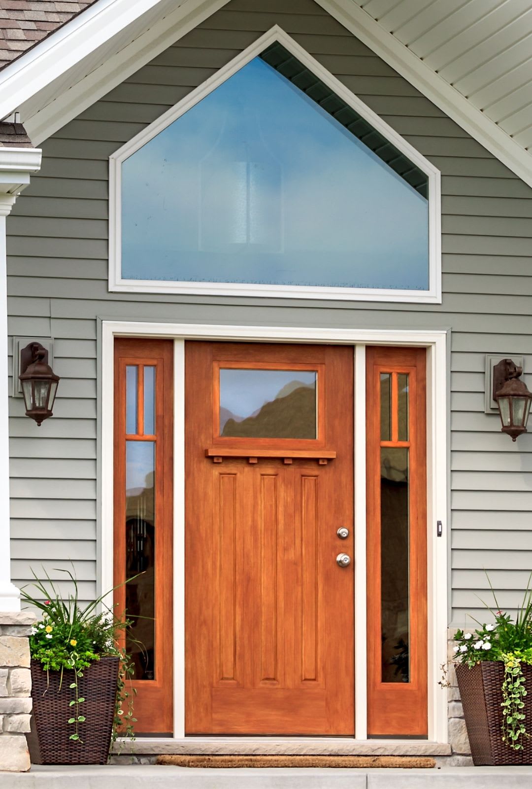 A wooden front door with glass panels, flanked by two potted plants, is set in a gray house with white trim and a large triangular window above, showcasing the elegance entry doors Chicago homes from Chicago Window Guys can offer.