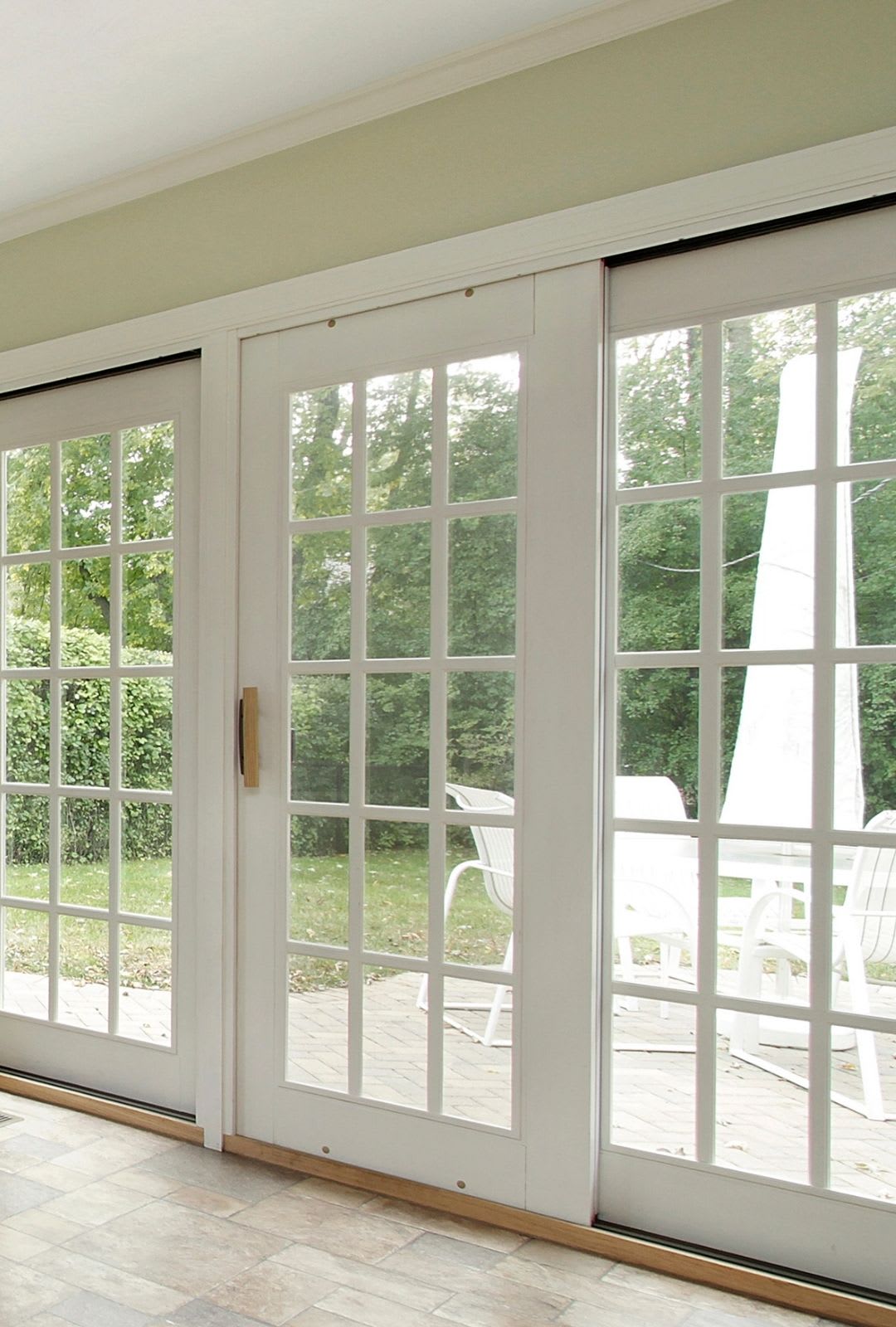 Large glass patio French doors with white frames open to a patio with white outdoor furniture and green trees in the background.