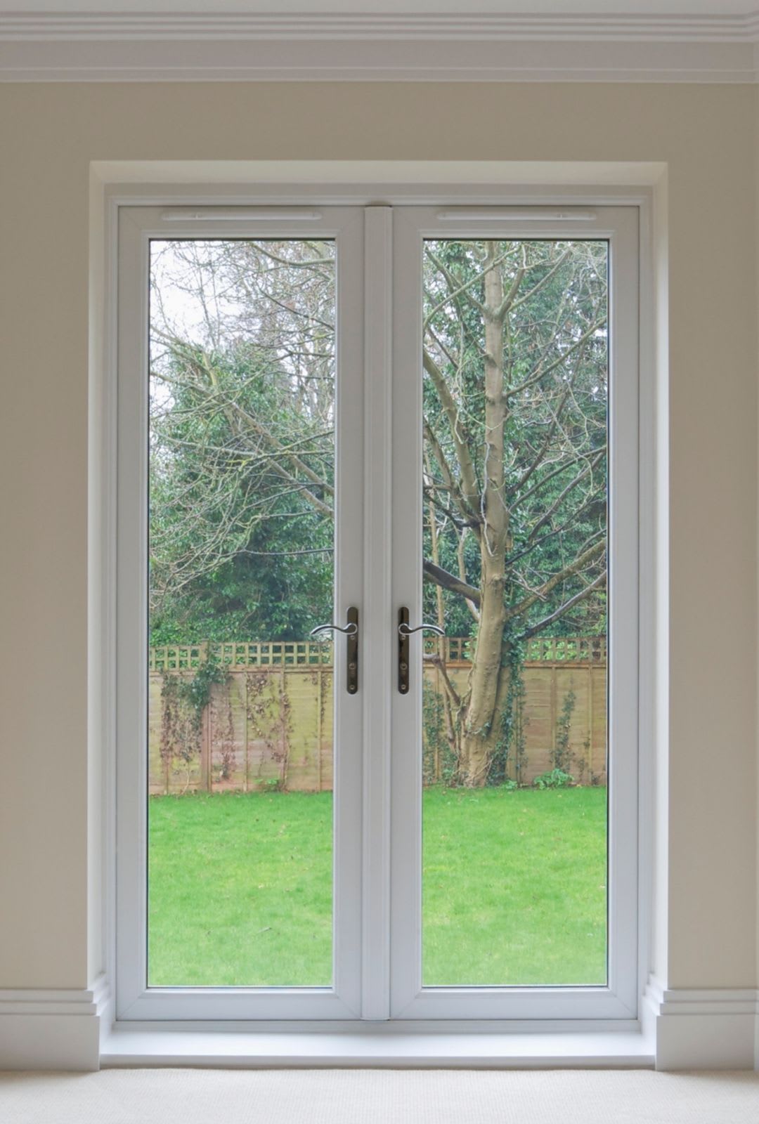 A pair of closed patio French doors in a beige room, looking out onto a green lawn, wooden fence, and trees in a garden—ideal for those considering patio door replacement to enhance their view and space.