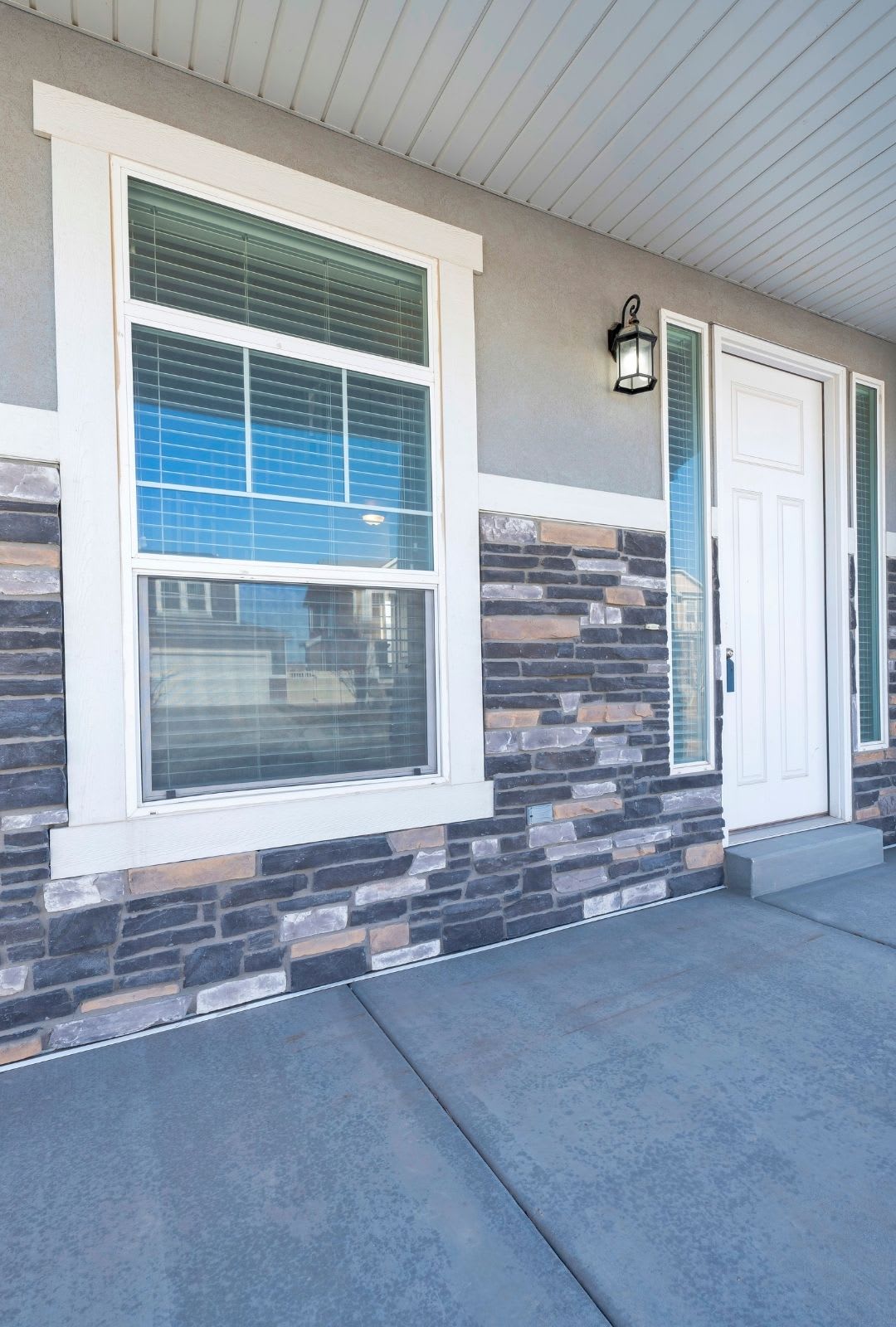 A front door and window of a house with stone and stucco exterior, white trim, concrete porch, outdoor wall light fixture, and stylish double-hung windows that enhance both charm and energy efficiency.