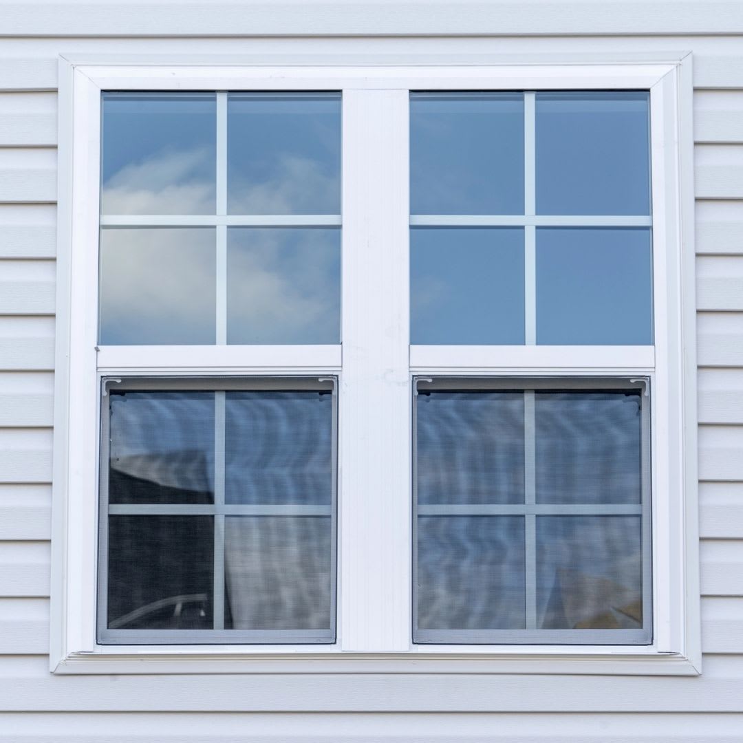 A double-hung window installation features white grid frames on a white-paneled exterior; the top panes reflect sky and clouds, while the bottom panes reveal interior curtains.