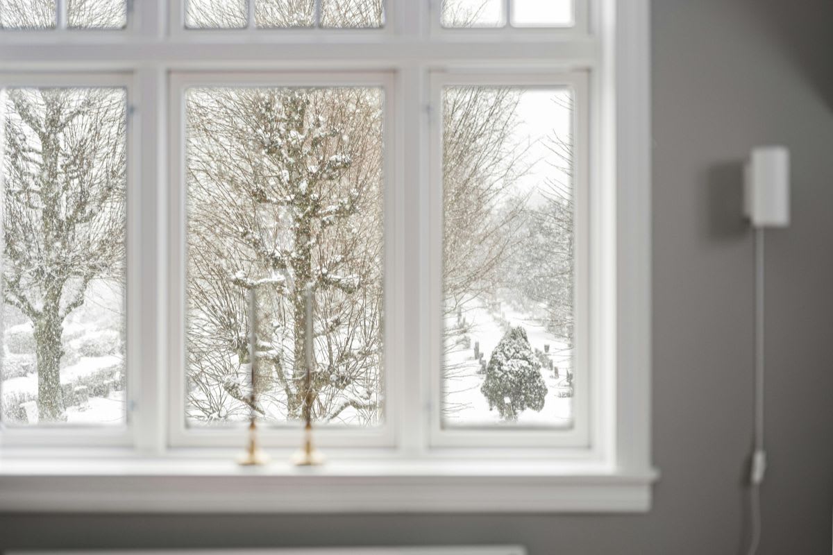 A snowy landscape with trees and bushes is visible through a multi-pane window in a gray-walled room, prompting the question: is it ok to replace windows in the winter?.