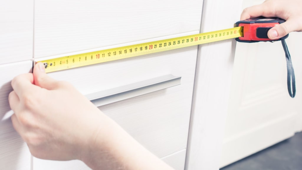 A person uses a tape measure to measure the width of a white cabinet drawer, much like taking door measurements when learning how to measure a door.