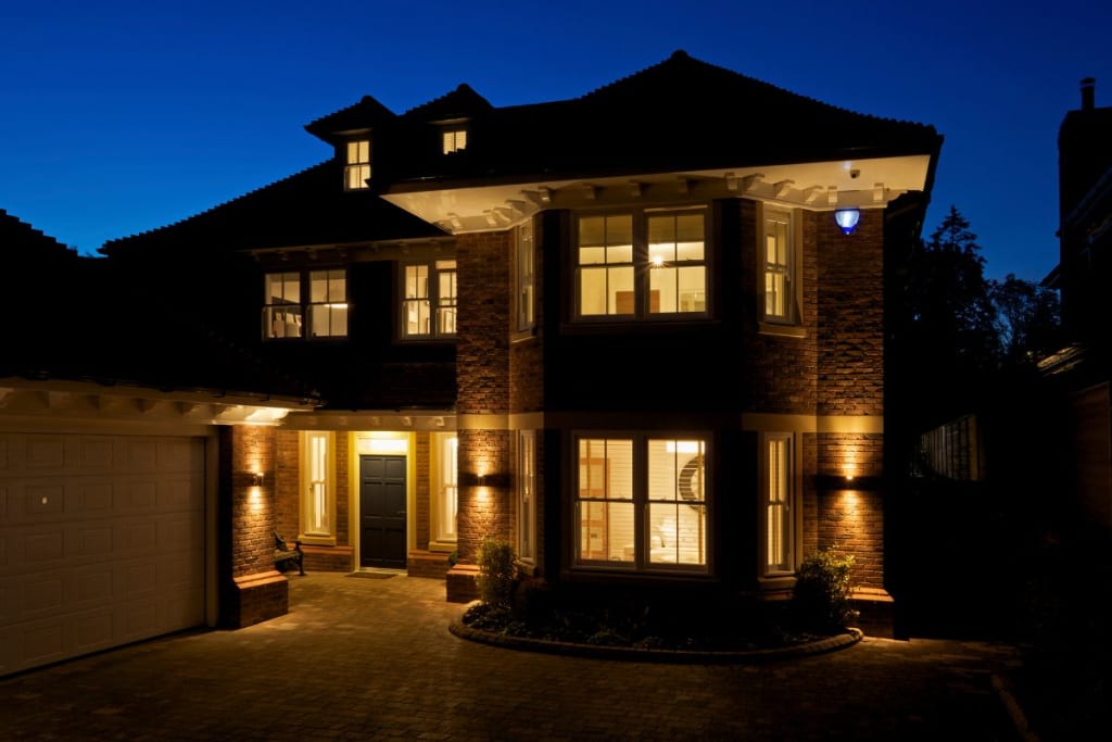 Two-story suburban house with burglar proof windows, warm exterior lighting, and a garage, photographed at dusk beneath a clear blue sky.