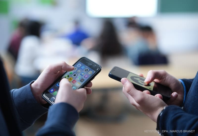 Zwei Schüler stehen in einem Klassenraum ab einem Gymnasium und schauen auf ihre Smartphones, auf einem der Displays ist das Logo der Social-Media-Plattform TikTok zu sehen.