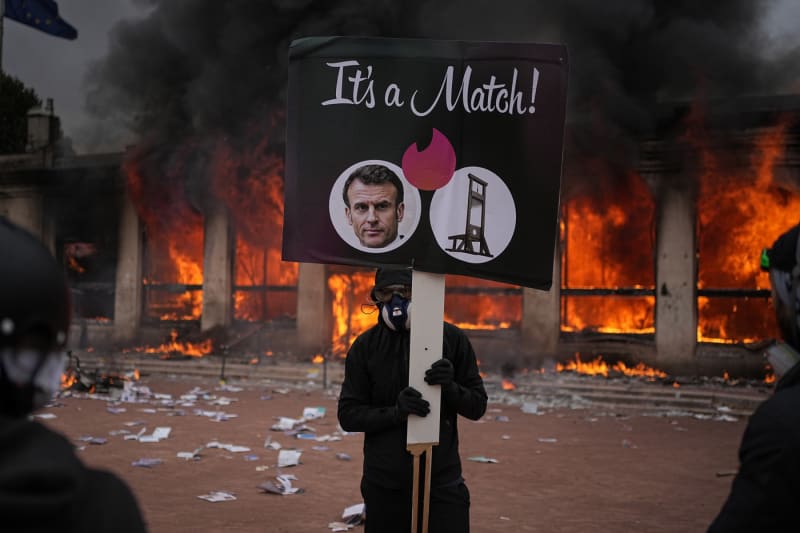 Ein Demonstrant in Lyon, Frankreich, hält ein satirisches Plakat hoch, auf dem der französische Präsident Emmanuel Macron und eine Guillotine im Stil der Dating-App Tinder mit dem Text 'It’s a Match!' zu sehen sind. Das geschah während der weit verbreiteten Demonstrationen und Ausschreitungen im Oktober 2026 wegen steigender Lebenshaltungskosten und Unzufriedenheit unter den Studenten.
