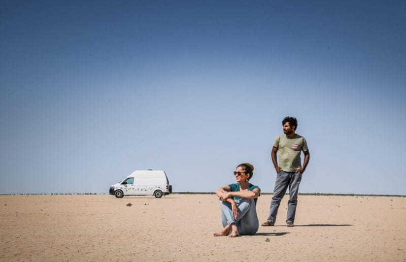 Martina und Dylan, die ganztägig in ihrem Wohnmobil leben, stehen in einer Wüste irgendwo auf ihrer Weltreise, während ihr Wohnmobil im Hintergrund zu sehen ist.