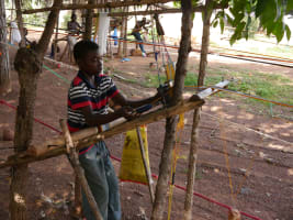 Mamadou steht an einem einfachen Holzwebstuhl im Freien und arbeitet mit bunten Fäden. Im Hintergrund sind weitere Kinder an Webstühlen zu sehen. Die Webstühle bestehen aus Holzstäben und vielen gespannten Fäden. Die Szene befindet sich unter Bäumen auf einem unbefestigten Boden.