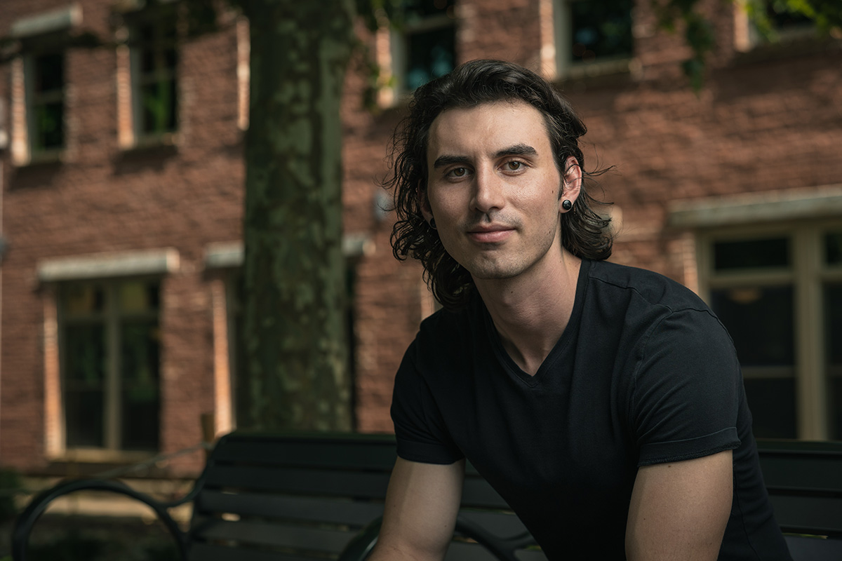 Young man wearing a black shirt sitting on a bench in downtown Asheville in front of a brick building for a headshot photography session.