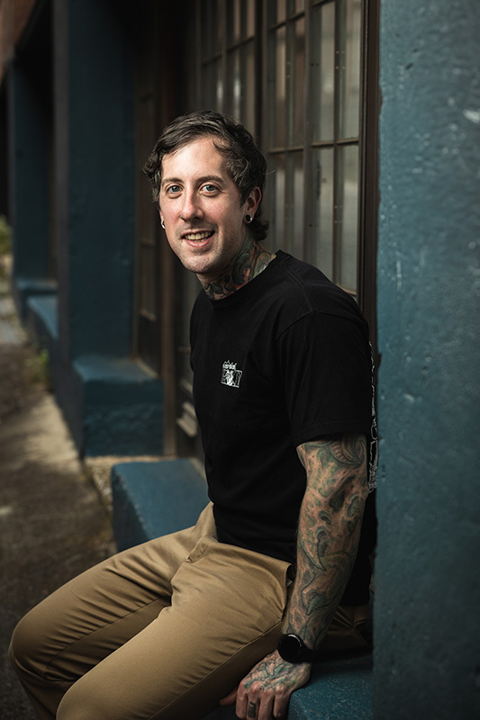 Tattooed Male wearing a black shirt and tan pants sitting on a dark green window seal.